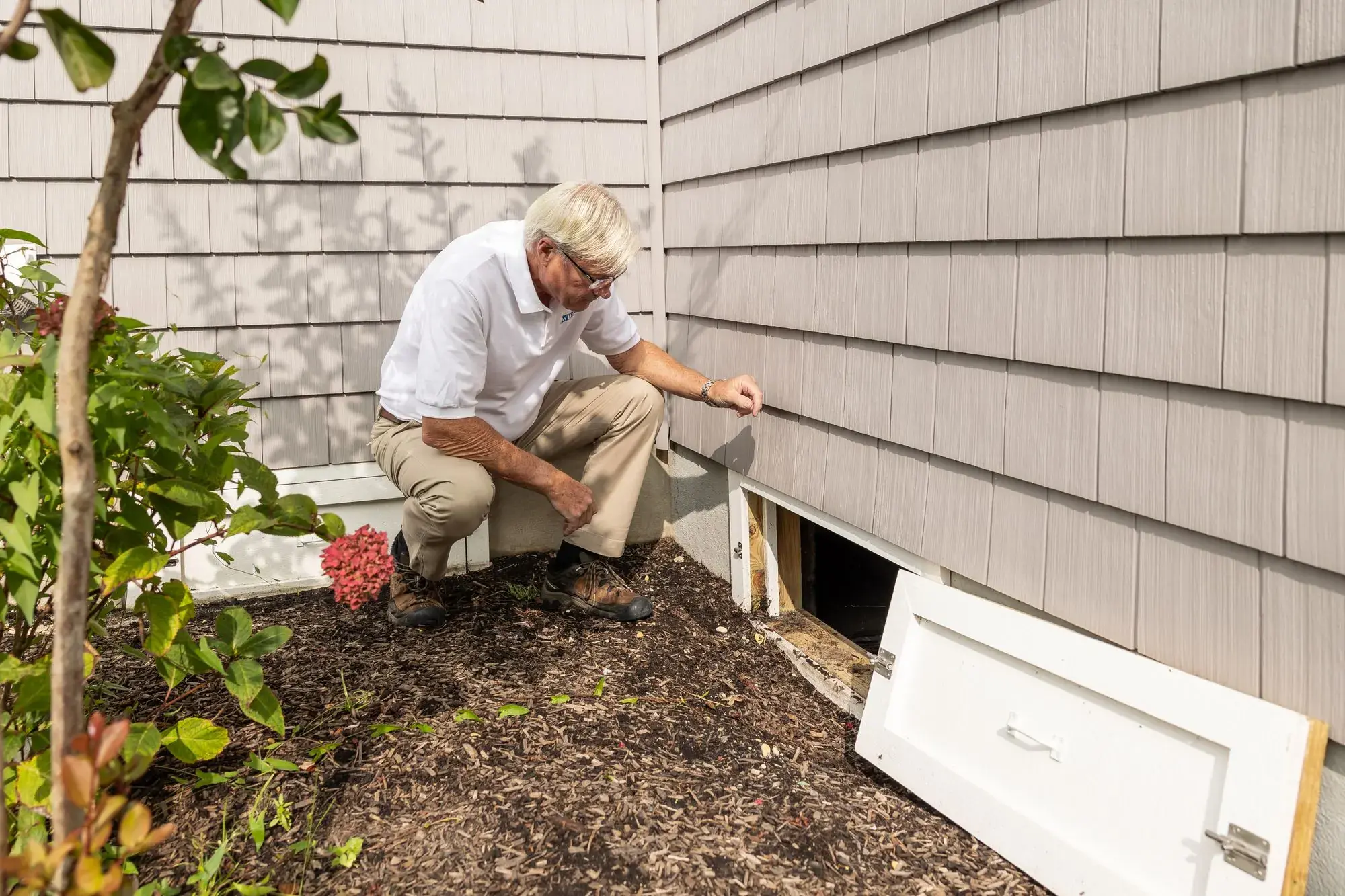Home inspector kneeling at a crawl space access door beside a house